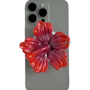 Peut inclure: Un support de téléphone en forme de fleur d'hibiscus rouge et rose, fixé à un smartphone gris. La fleur a des pétales détaillés et une finition brillante. Le téléphone a un appareil photo à trois objectifs.