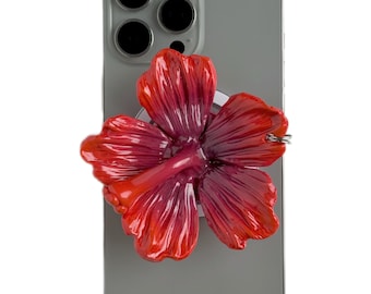 May include: A red and pink hibiscus flower phone grip attached to a gray smartphone. The flower has detailed petals and a glossy finish. The phone has a triple-lens camera.
