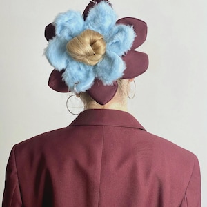 Peut inclure: Un blazer bordeaux avec un grand couvre-chef en forme de fleur. Le couvre-chef présente des pétales bordeaux, des accents duveteux bleu clair et un centre de cheveux blonds. Des créoles argentées sont visibles.