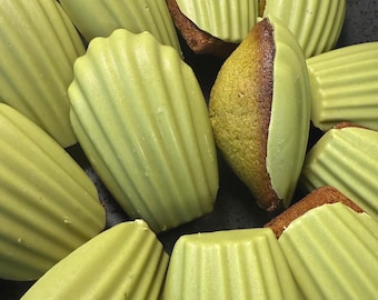 May include: A close-up shot of several madeleine cookies arranged on a dark plate. The cookies have a light green, glossy coating and a distinctive shell shape. Some cookies are cut open, revealing a brown interior.