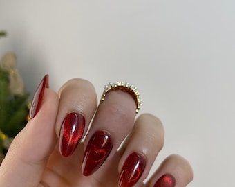 May include: Close-up of a hand with red almond-shaped nails with a subtle heart design. The nails are painted with a red shimmer polish.