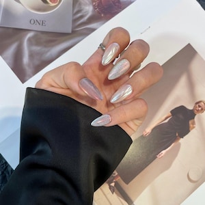 May include: Close-up of a hand with long, almond-shaped nails painted with a variety of iridescent and glitter nail polish.