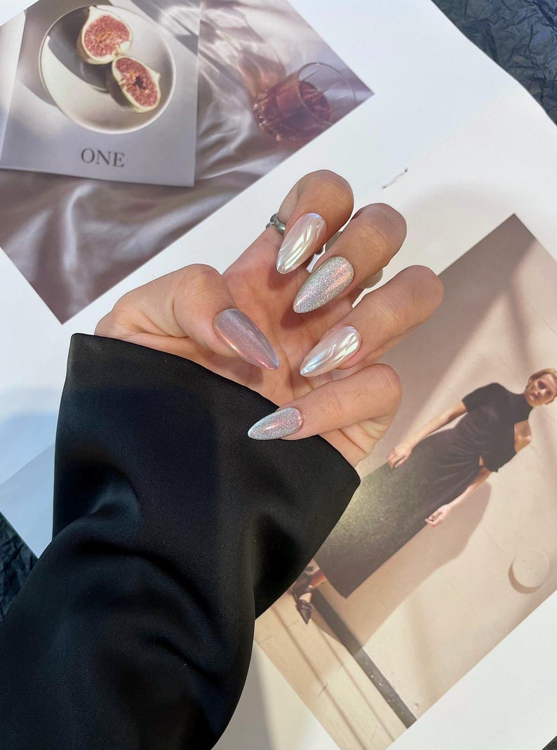 May include: Close-up of a hand with long, almond-shaped nails painted with a variety of iridescent and glitter nail polish.