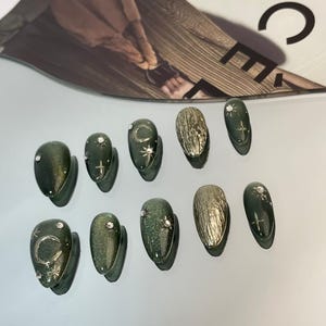 May include: A set of ten artificial nails in various designs. Some nails are a dark green with glitter, adorned with silver crescent moons, stars, and rhinestones. Other nails have a textured gold finish. The nails are oval-shaped.