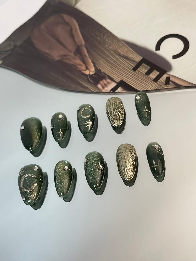 May include: A set of ten artificial nails in various designs. Some nails are a dark green with glitter, adorned with silver crescent moons, stars, and rhinestones. Other nails have a textured gold finish. The nails are oval-shaped.