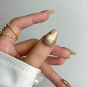 May include: Close-up of a hand with long, almond-shaped gold nails. One nail features a gold star design. A gold ring adorns a finger. The nails have a glittery finish, and the hand is against a white background.