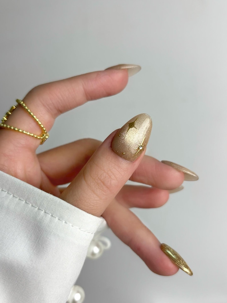 May include: Close-up of a hand with long, almond-shaped gold nails. One nail features a gold star design. A gold ring adorns a finger. The nails have a glittery finish, and the hand is against a white background.