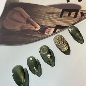May include: A set of six dark green, almond-shaped artificial nails. Some nails feature silver embellishments, including a small gem, a crescent moon, and a starburst design. One nail has a textured gold surface.