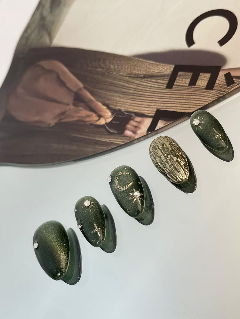 May include: A set of six dark green, almond-shaped artificial nails. Some nails feature silver embellishments, including a small gem, a crescent moon, and a starburst design. One nail has a textured gold surface.