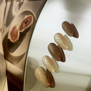 May include: A set of six oval-shaped nail polish colors. The colors are a light brown with gold glitter, a light brown with a swirl pattern, a light brown with gold glitter, a light brown with a swirl pattern, a light brown with gold glitter, and a light brown with a swirl pattern.