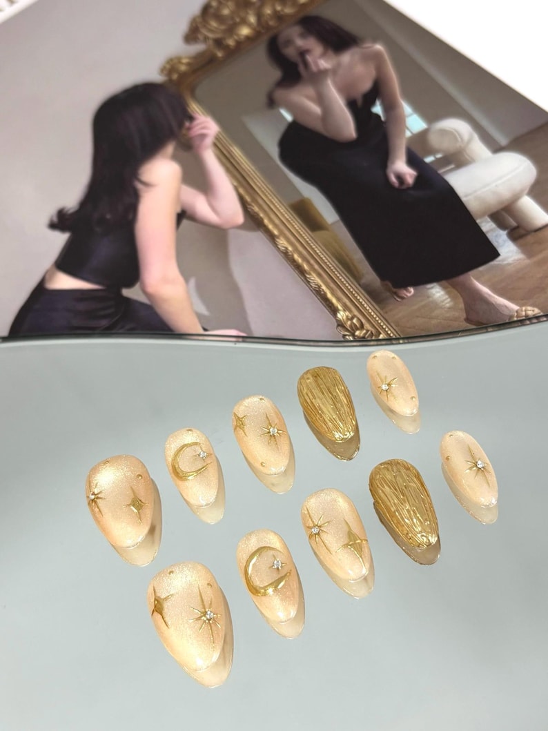 May include: A set of ten almond-shaped artificial nails. The nails are a light beige with gold accents. Some nails feature a gold crescent moon and star design, while others have a textured gold finish. Small rhinestones add sparkle.