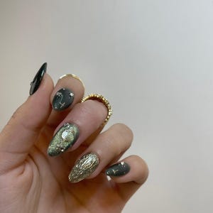 Könnte beinhalten: Eine Hand mit langen, oval geformten, manikürten Nägeln. Die Nägel weisen eine Vielzahl von Designs auf, darunter eine dunkelgrüne Basis mit silbernem Glitzer, Goldfolie und klaren Edelsteinen. Goldringe schmücken die Finger.