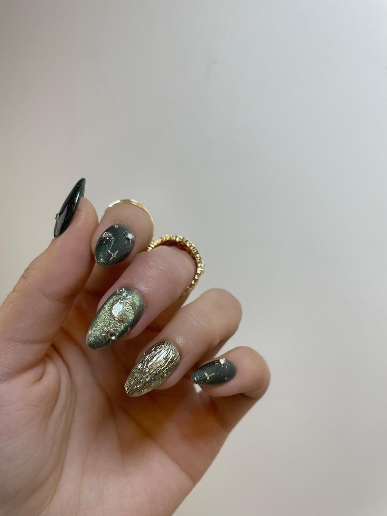 May include: A hand with long, oval-shaped, manicured nails. The nails feature a variety of designs, including a dark green base with silver glitter, gold foil, and clear gemstones. Gold rings adorn the fingers.