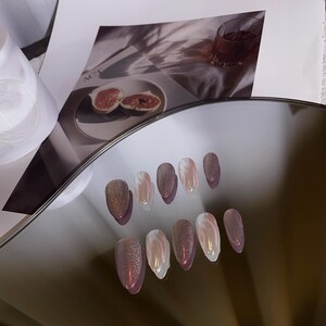 May include: A set of ten false nails with a shimmery, iridescent finish. The nails are a light pink color with a white swirl design. The nails are arranged in two rows on a reflective surface.