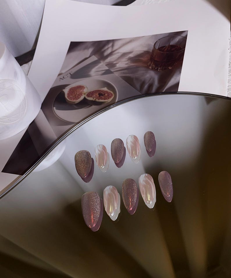May include: A set of ten false nails with a shimmery, iridescent finish. The nails are a light pink color with a white swirl design. The nails are arranged in two rows on a reflective surface.
