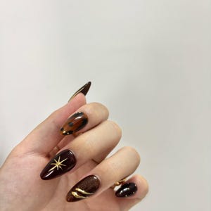 Może przedstawiać: Dłoń prezentująca migdałowe, ciemnobrązowe sztuczne paznokcie. Paznokcie zdobione są złotymi akcentami, w tym wzorem gwiazdy i zakrzywionymi liniami. Jeden paznokieć ma wzór szylkretu.