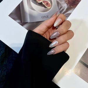 May include: Close-up of a hand with long, almond-shaped nails. The nails are painted with a silver glitter polish and a white and silver iridescent polish. The nails are decorated with a swirl design.
