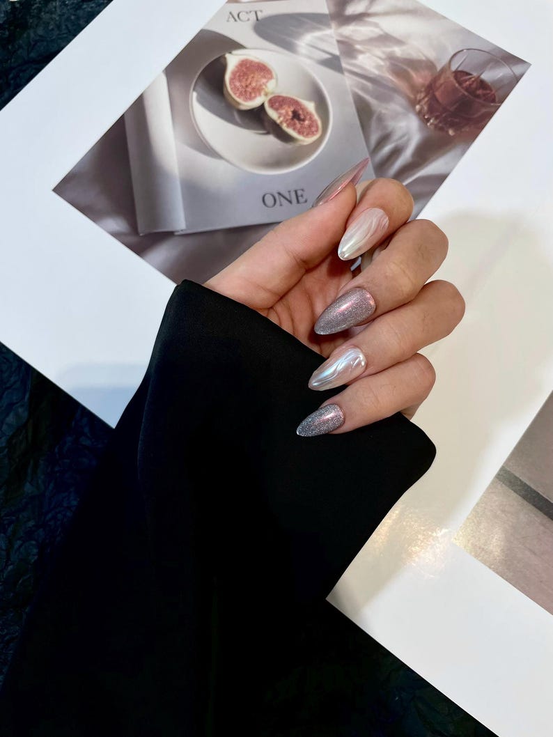 May include: Close-up of a hand with long, almond-shaped nails. The nails are painted with a silver glitter polish and a white and silver iridescent polish. The nails are decorated with a swirl design.