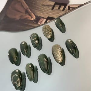 May include: A collection of artificial nails in various designs. The nails feature a dark green glitter finish, gold textured accents, and silver embellishments. Some nails have crescent moon and star designs. The nails are arranged on a light gray surface.