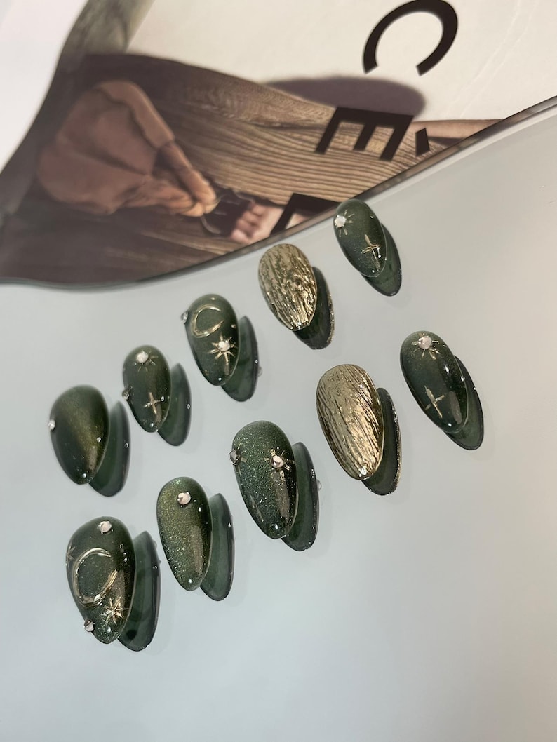May include: A collection of artificial nails in various designs. The nails feature a dark green glitter finish, gold textured accents, and silver embellishments. Some nails have crescent moon and star designs. The nails are arranged on a light gray surface.