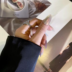 May include: A silver ring with a unique abstract design. The ring is worn on the index finger of a hand with long, pointed nails painted with a shimmery, iridescent polish.