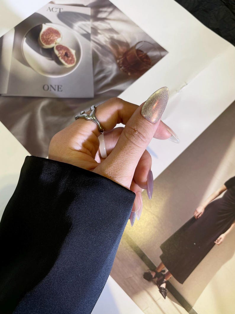 May include: A silver ring with a unique abstract design. The ring is worn on the index finger of a hand with long, pointed nails painted with a shimmery, iridescent polish.