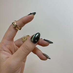May include: Hand with gold rings and long, dark green nails. One nail has a small, clear gem. The rings include a wavy gold band and a textured gold ring with pearls. The nails have a glossy finish.