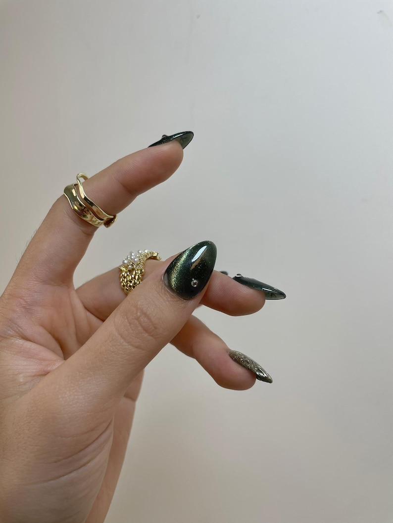 May include: Hand with gold rings and long, dark green nails. One nail has a small, clear gem. The rings include a wavy gold band and a textured gold ring with pearls. The nails have a glossy finish.