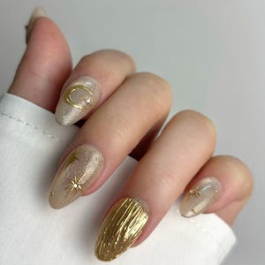 May include: A hand with almond-shaped, gold-toned nails. Some nails feature gold crescent moons, stars, and textured gold designs. The nails have a glittery, shimmering finish, with a neutral base color.