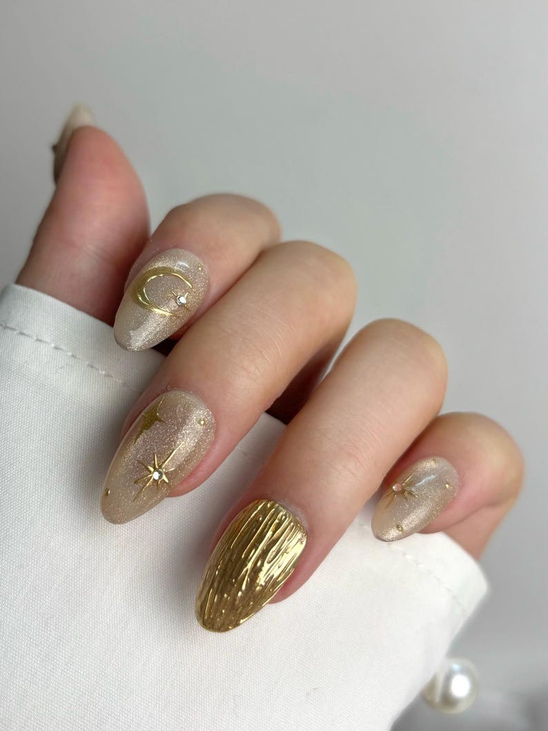 May include: A hand with almond-shaped, gold-toned nails. Some nails feature gold crescent moons, stars, and textured gold designs. The nails have a glittery, shimmering finish, with a neutral base color.