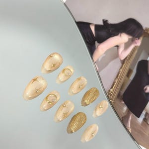 May include: A set of ten artificial nails with a light beige base and gold accents. Designs include stars, moons, and textured gold elements. The nails are arranged on a reflective surface.