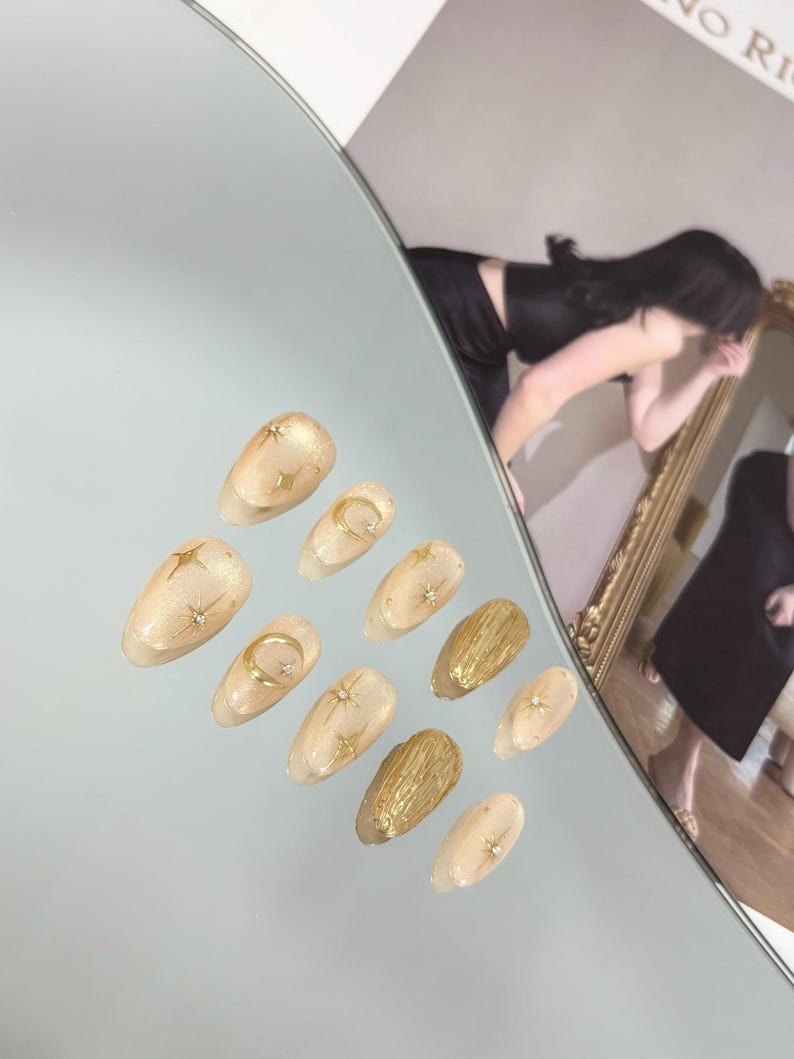 May include: A set of ten artificial nails with a light beige base and gold accents. Designs include stars, moons, and textured gold elements. The nails are arranged on a reflective surface.