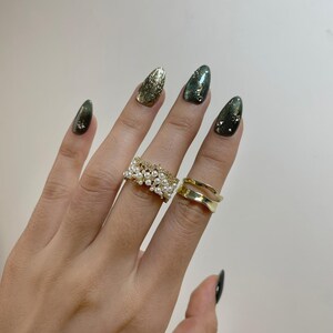 May include: A hand wearing two gold rings and with long, almond-shaped nails painted in shades of green and gold. One ring is a wide band with small pearls, and the other is a double-band design.