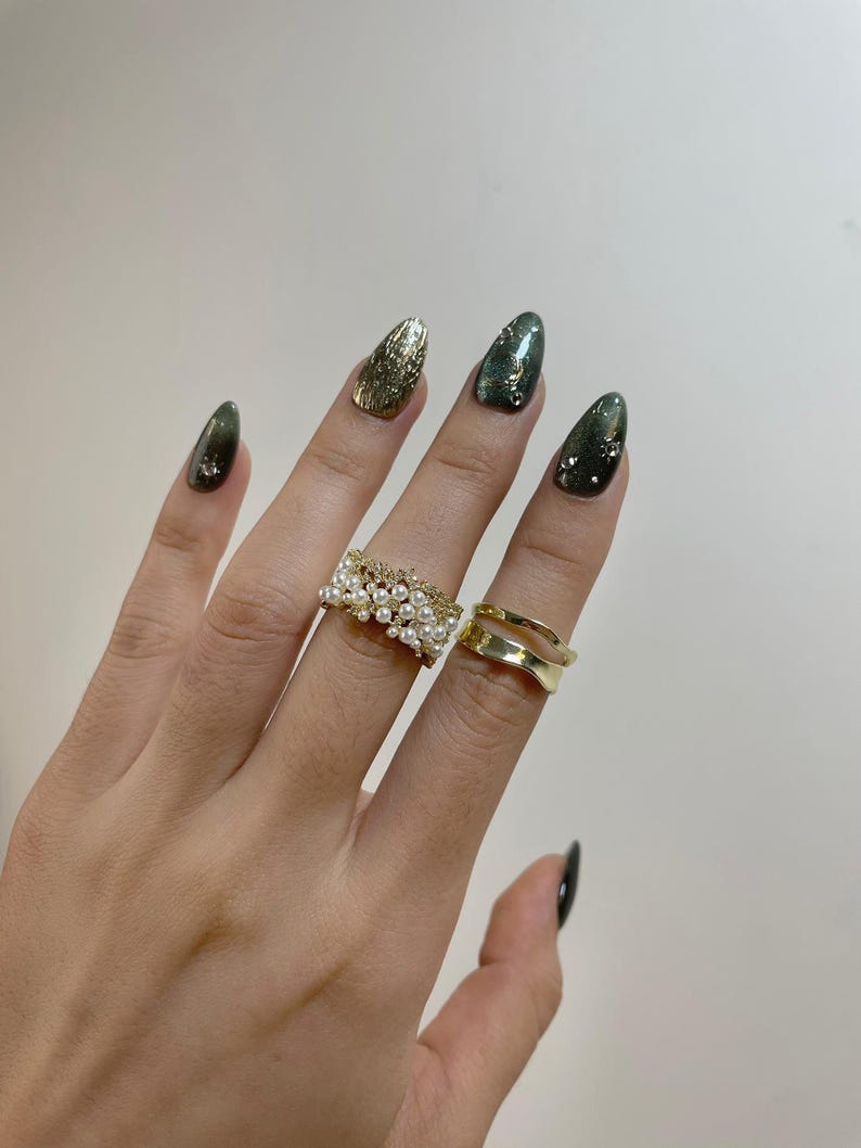 May include: A hand wearing two gold rings and with long, almond-shaped nails painted in shades of green and gold. One ring is a wide band with small pearls, and the other is a double-band design.