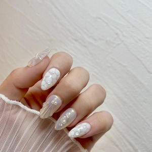 Könnte beinhalten: Weiße ovale Nägel mit Perlenakzenten. Einige Nägel haben ein strukturiertes Design, während andere ein Muscheldesign haben.