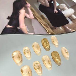 May include: A set of ten almond-shaped artificial nails. The nails are a light beige with gold star and crescent moon designs. Some nails have a textured gold design. Small rhinestones are placed on some nails.