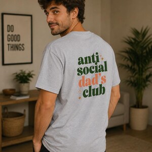 Peut inclure: T-shirt gris clair avec l'inscription "anti social dad's club" en vert et pêche. Le t-shirt est de style col rond et semble être fait d'une matière douce. Une personne porte le t-shirt.