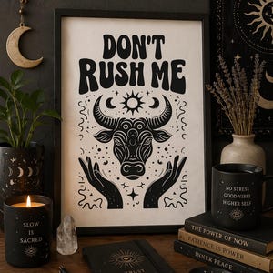 Könnte beinhalten: Ein gerahmter Druck mit den Worten "DON'T RUSH ME" über einer Stierillustration. Eine schwarze Kerze mit dem Text "SLOW IS SACRED", eine schwarze Tasse mit dem Text "NO STRESS GOOD VIBES HIGHER SELF" und ein Notizbuch mit dem Text "TRUST THE PROCESS" sind ebenfalls zu sehen.