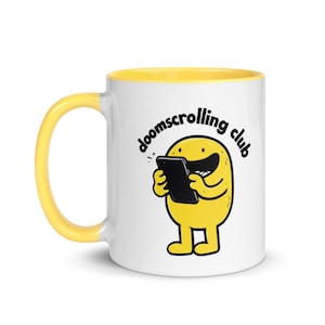 Peut inclure: Mug en céramique blanche avec intérieur et anse jaunes. Le mug présente un personnage de dessin animé jaune tenant un smartphone noir, avec l'inscription "doomscrolling club" en forme incurvée.