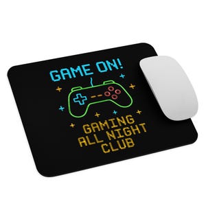 May include: Black rectangular mousepad with the text "GAME ON!" in blue, a green and red video game controller graphic, and the text "GAMING ALL NIGHT CLUB" in yellow. A white mouse is next to the mousepad.