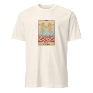 Peut inclure: Un t-shirt à manches courtes blanc cassé avec un motif rectangulaire représentant deux silhouettes se tenant la main au-dessus de vagues stylisées roses et rouges. Le texte GEMINI figure en haut et DEPENDS ON THE MOOD en bas.