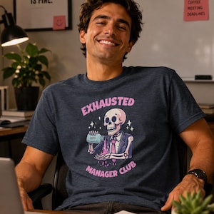 Peut inclure: T-shirt bleu marine avec l'inscription "EXHAUSTED MANAGER CLUB" et un dessin de squelette en costume et cravate, tenant une bouteille. La personne sur la photo sourit. L'arrière-plan comprend des éléments de bureau.