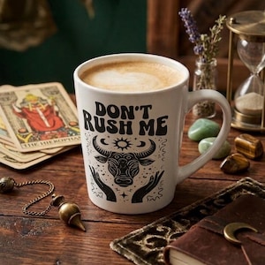 Peut inclure: Une tasse à café blanche avec le texte "DON'T RUSH ME" et un motif de taureau, remplie de latte. La tasse est posée sur une table en bois avec des cartes de tarot, des cristaux et un journal en cuir. Un sablier et des fleurs séchées sont en arrière-plan.