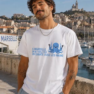 Puede incluir: Camiseta blanca con un pulpo azul y el texto "A Marseille, on ne parle pas, on chante avec les mains." La camiseta es de un material suave y es adecuada para uso casual. La camiseta la lleva una persona.