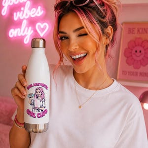 Könnte beinhalten: Eine weiße Edelstahl-Trinkflasche mit silbernem Deckel. Die Flasche zeigt eine rosa-lila Grafik einer Frau, die ein Getränk hält, mit dem Text "Nova Aesthetic Girl Club". Der Hintergrund enthält ein pinkfarbenes Neon-Schild mit der Aufschrift "good vibes only".
