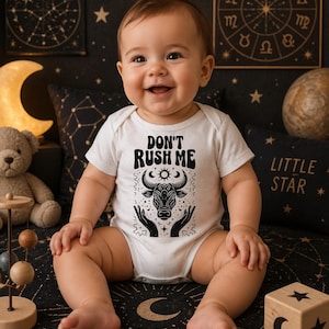 Könnte beinhalten: Ein lächelndes Baby trägt einen weißen Body mit dem Text "DON'T RUSH ME" und einer schwarzen Grafik eines Stierkopfes. Das Baby sitzt auf einer schwarzen, gemusterten Oberfläche mit Himmelsdesigns, umgeben von dekorativen Kissen und Spielzeug.