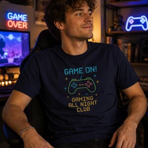 May include: Navy blue t-shirt with the text "GAME ON!" and a neon green and blue controller graphic. The shirt also says "GAMING ALL NIGHT CLUB" in a pixelated font. The person is holding a black game controller.