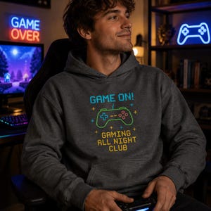 May include: A dark gray hoodie with the text "GAME ON!" and "GAMING ALL NIGHT CLUB" in a pixelated font. The design includes a green and blue controller graphic. The person is holding a black controller.