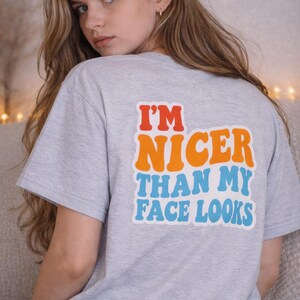 Peut inclure: Un t-shirt gris clair avec le texte "I'M NICER THAN MY FACE LOOKS" en orange, bleu et rouge. Le t-shirt à manches courtes est porté par une personne aux longs cheveux blonds.