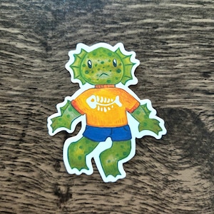 May include: A sticker featuring a cartoon creature with green skin and a fish-bone design on its orange shirt. The creature has blue shorts and a serious expression. The sticker is cut around the edges of the illustration.